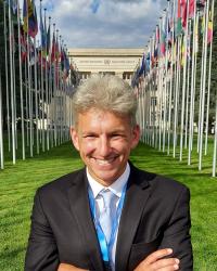 Photo of middle-aged white man with white hair, blue eyes, and smiling dressed in white shirt, blue tie, and black blazer standing in front of the United Nations building in Geneva.