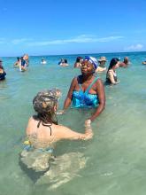Image of a Jamaican women in a blue swimsuit holding hands with student in the ocean shore
