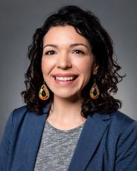 Portrait of Theresa Rocha Beardall with dark curly hair, blue blazer, and charcoal shirt. 