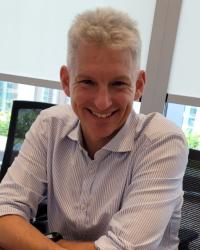Photo of a middle-aged white man with white hair, blue eyes, and clean-shaven face sitting at a desk.