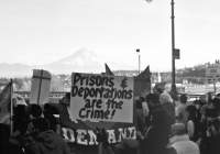 Black and white photo of protestors. In the center is a sign or poster that says "Prisons& deportations are the crime!"