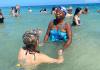 Image of a Jamaican women in a blue swimsuit holding hands with student in the ocean shore